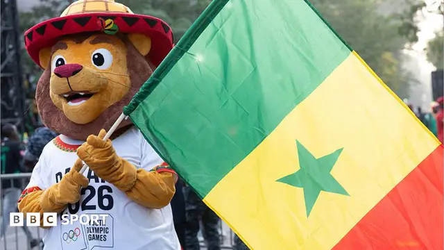 A friendly-looking lion mascot holding up a large national flag of Senegal on a pole. The mascot, called Ayo, wears a colourful red-and-yellow traditional-style hat and a white T-shirt featuring the logo for the Dakar 20