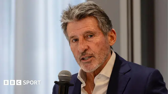 Sebastian Coe, visible from shoulders up wearing a white open-necked shirt and a navy blue suit jacket, holds a microphone up to his mouth as he speaks at a news conference. A wall and a window can be seen out of focus b