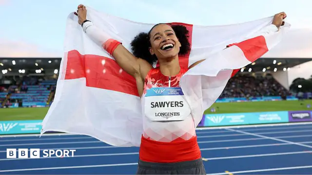 A smiling Jazmin Sawyers in a red and white Team England vest raises her arms to the skies whiles draped in the England flag