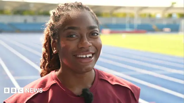 Success Eduan has long brown hair which is braided and tied back in a pony tail. She is wearing a dark red top and is standing on a blue running track while smiling for the camera.